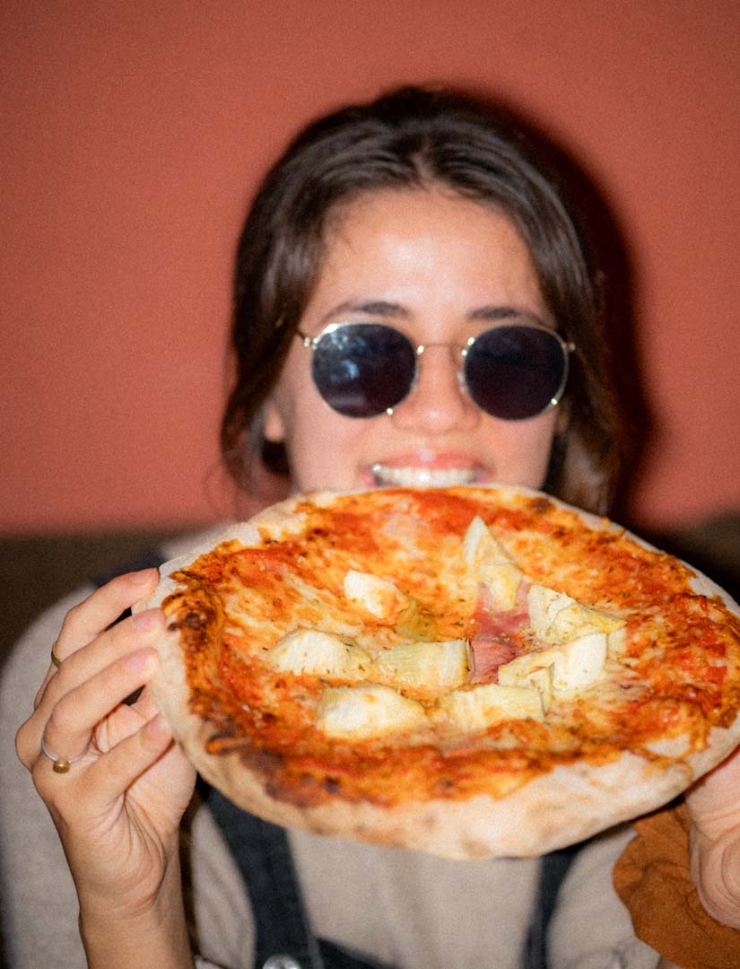 Eine Studentin beißt mit einem glücklichen Gesichtsausdruck in eine noch nicht angeschnittene Pizza.