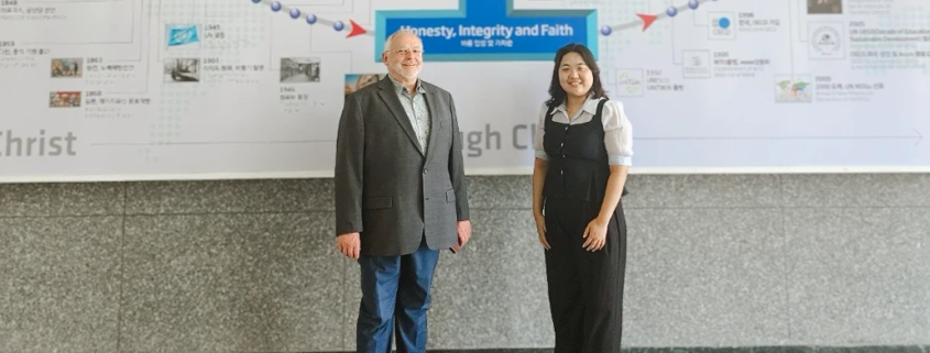 Dr. Friedemann Burkhardt steht gemeinsam mit einer Frau vor einer Wandgrafik an der Handong Global University in Südkorea. Auf der Grafik sind die Worte ‚Honesty, Integrity and Faith‘ zu lesen