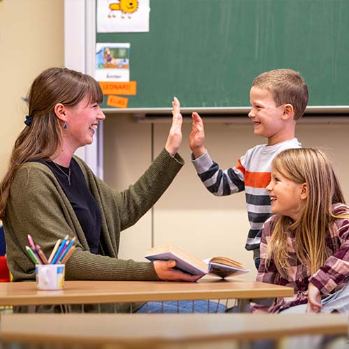 Eine Studentin sitzt mit zwei Grundschulkindern im Klassenzimmer, liest aus einem Buch vor und klatscht mit einem Jungen ein. Ein Mädchen neben ihm lächelt