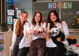 3 Studentinnen sitzen lächelnd im Klassenzimmer einer Grundschule und halten die Buchstaben IHL in die Kamera