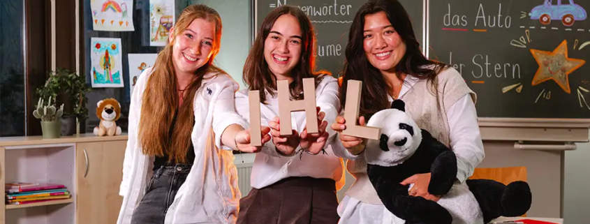 3 Studentinnen sitzen lächelnd im Klassenzimmer einer Grundschule und halten die Buchstaben IHL in die Kamera