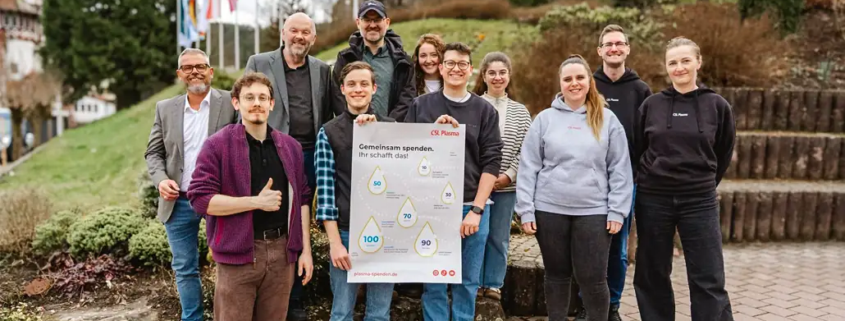 „Eine Gruppe Studierender steht gemeinsam mit Vertretern der Hochschule, der Liebenzeller Mission und Mitarbeitenden von CSL Plasma für ein Gruppenfoto. Sie halten ein Plakat mit der Aufschrift ‚Gemeinsam spenden. Ihr schafft das.‘.“